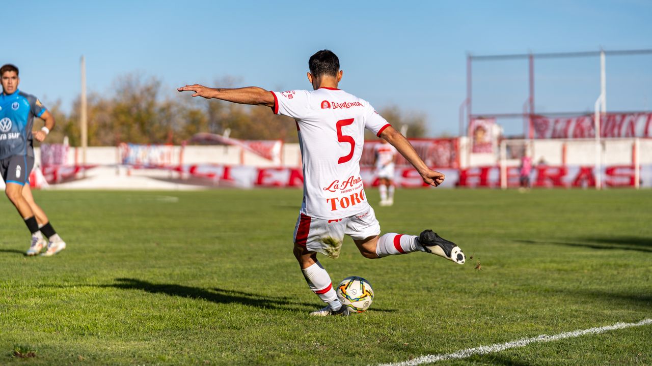 Soccer player in white jersey taking a kick during a competitive match under bright daylight on a grassy field.